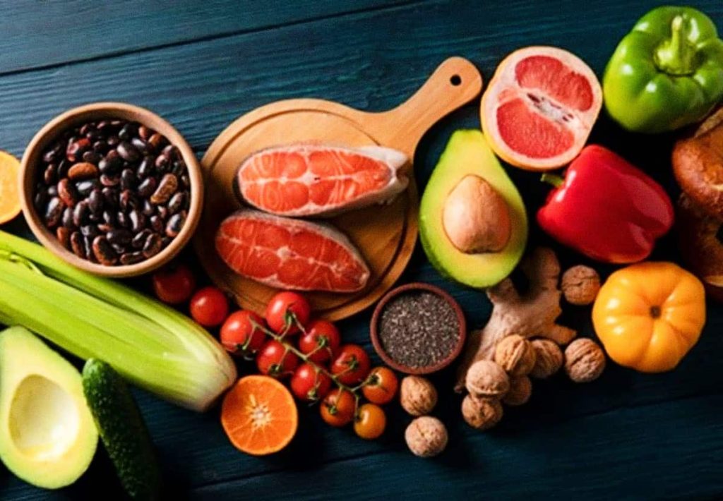 A top-down view of a colorful assortment of whole foods including salmon steaks, avocado, leafy greens, legumes, bell peppers, and citrus, representing a complete potassium-rich diet.