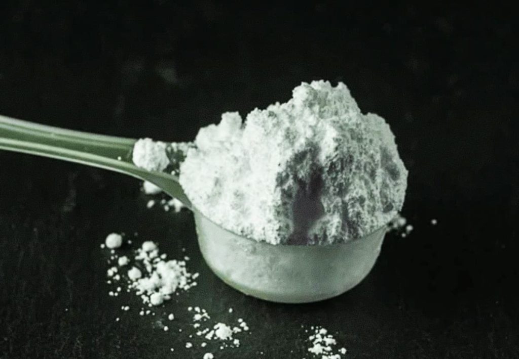 A macro close-up of a white supplement scoop filled with pure micronized creatine powder on a dark background.