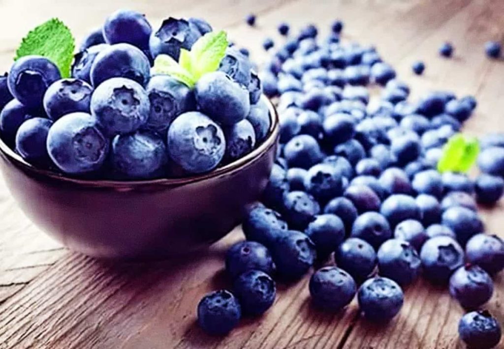 A dark bowl filled with fresh blueberries and mint leaves, with more berries scattered on a wooden surface.
