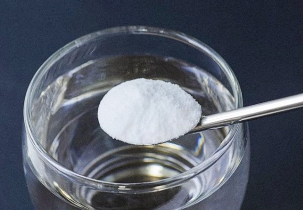 A metal spoon holding white creatine powder over a clear glass of water, about to be mixed.
