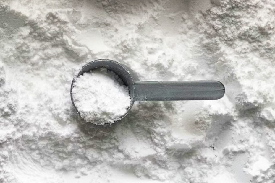 A top-down view of a grey plastic scoop filled with white creatine monohydrate powder, resting on a bed of the same powder.