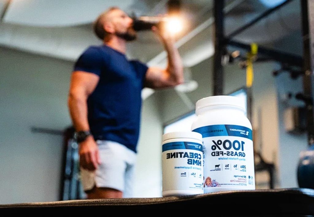 A muscular man in a gym setting drinking from a shaker bottle, with containers of Creatine and 100% Grass-Fed Whey Protein in the foreground.