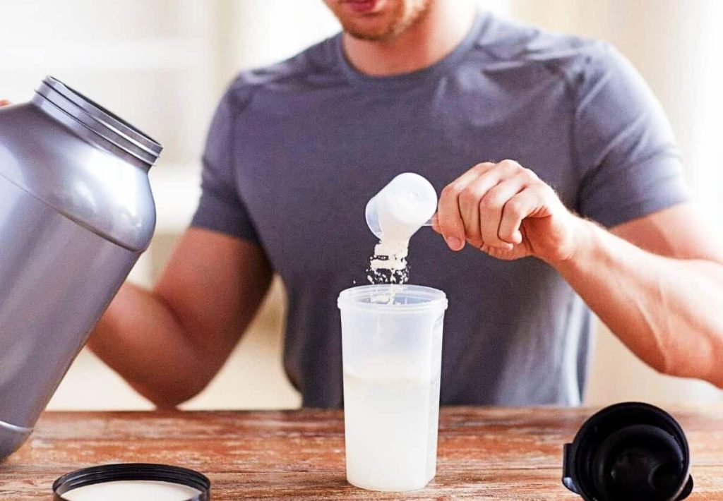 A close-up of a man pouring a white supplement scoop into a shaker bottle.