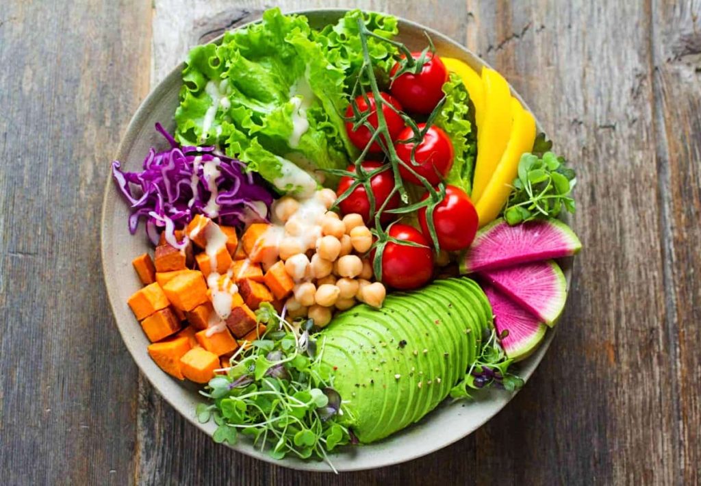 A vibrant Buddha bowl on a wooden table containing avocado, sweet potatoes, chickpeas, cherry tomatoes, lettuce, and watermelon radish.