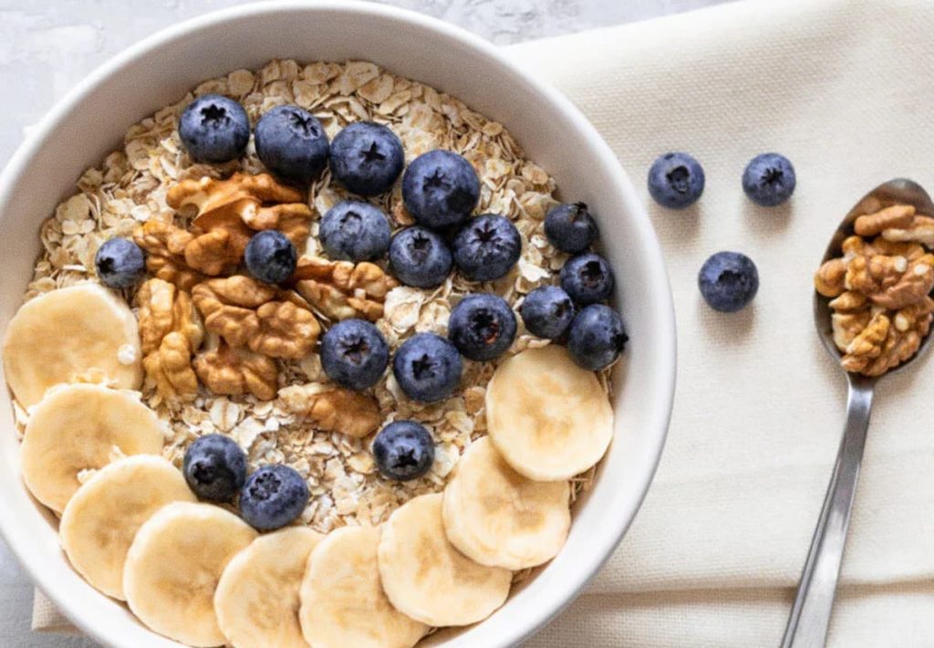 A white bowl of oatmeal topped with fresh blueberries, walnuts, and sliced bananas, illustrating slow-releasing complex carbohydrates.