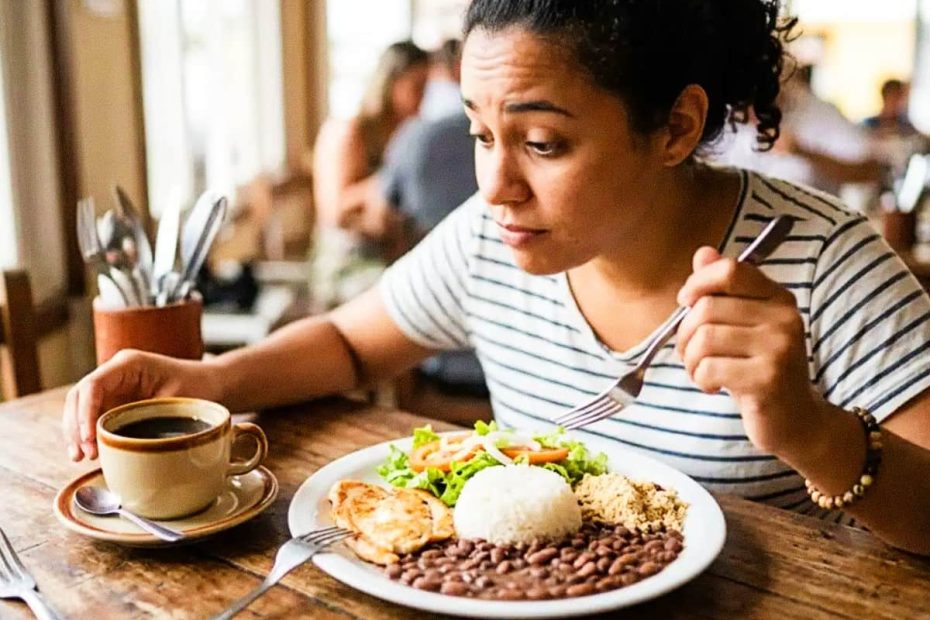 A woman at a wooden dining table looking at a cup of coffee while eating a traditional meal of rice, beans, and salad.