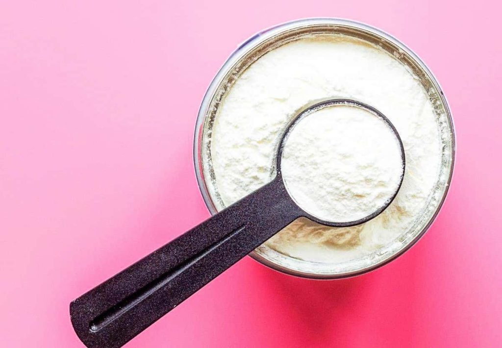 A top-down view of a black measuring scoop filled with white creatine powder inside a container on a bright pink background.