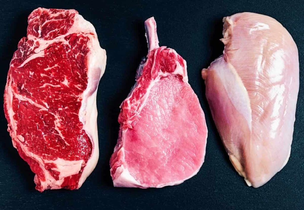 A side-by-side comparison of raw beef steak, pork loin, and chicken breast on a black background.