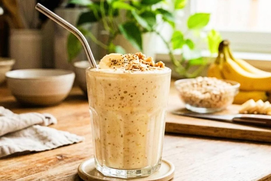 A creamy banana and oatmeal smoothie in a tall ribbed glass with a metal straw, topped with walnuts and oats on a wooden table.