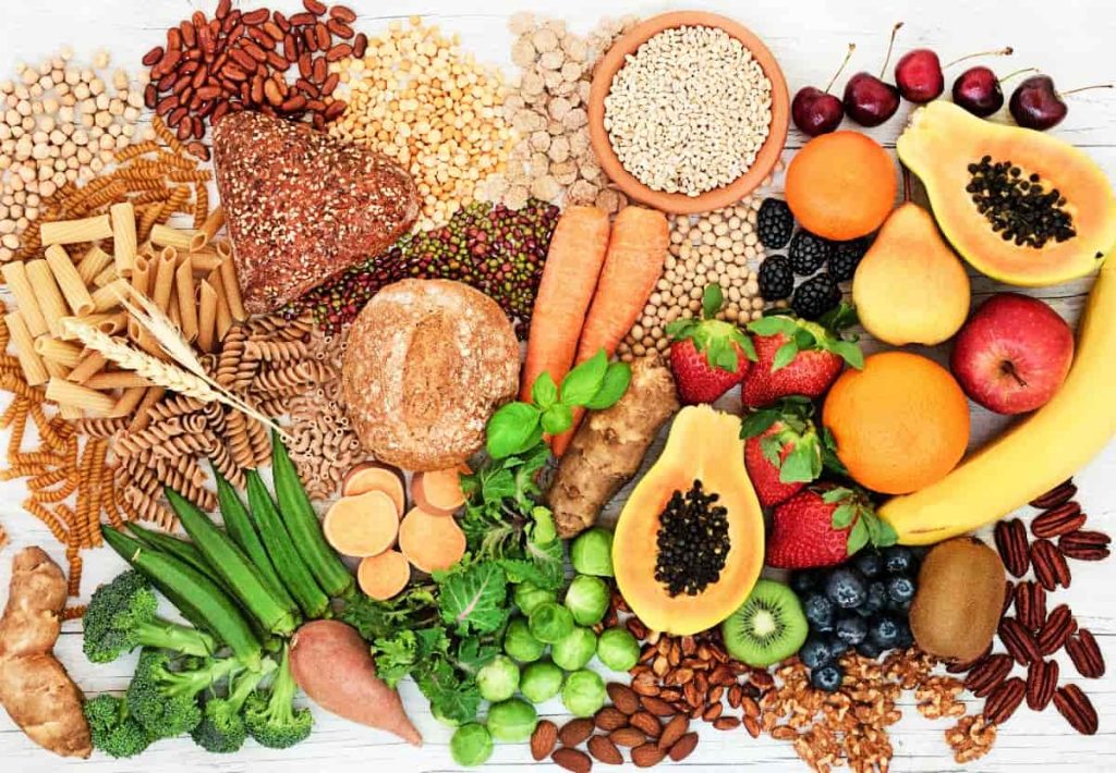 Overhead view of various healthy foods including broccoli, strawberries, whole grains, nuts, bananas, and papaya arranged on a light background.