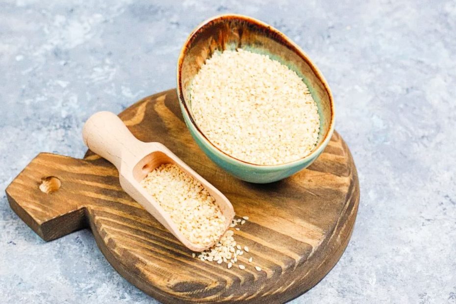 Whole psyllium seeds in a small green ceramic bowl and a wooden scoop on a rustic wooden board.