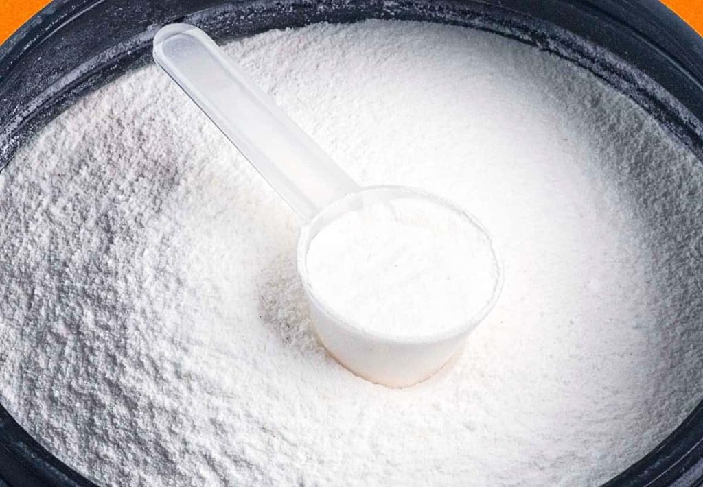 Person holding a single scoop of creatine powder over a blue supplement tub.