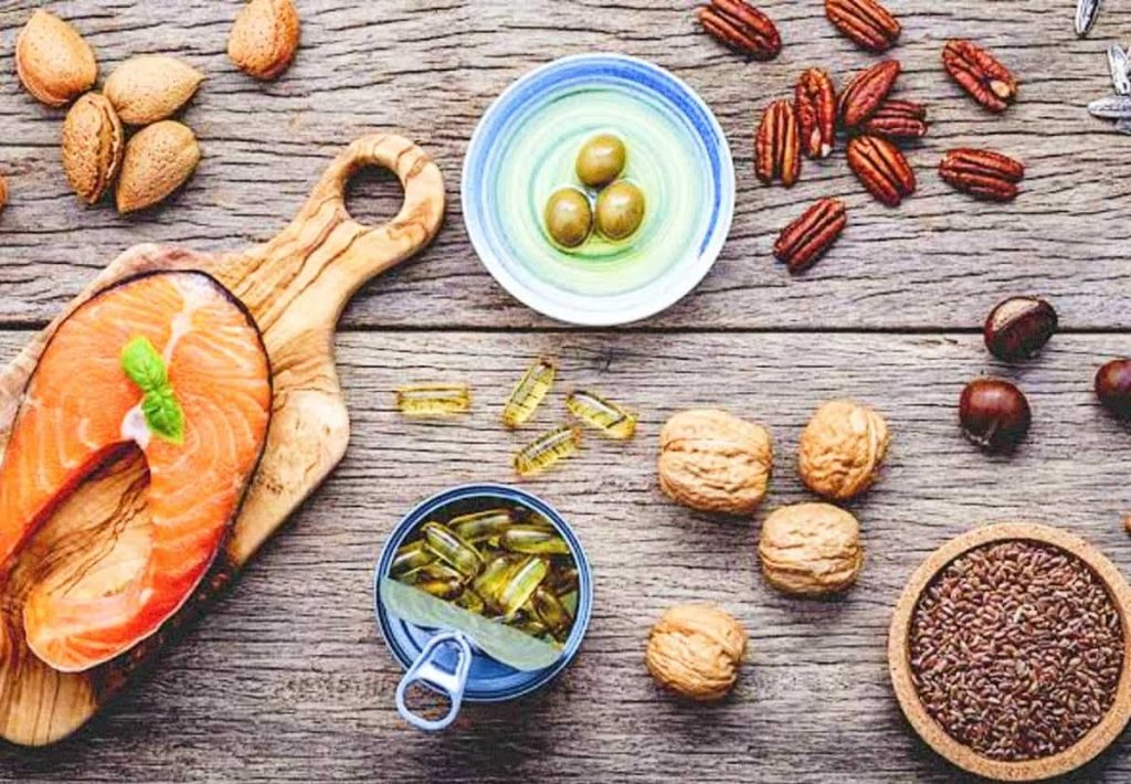 A rustic overhead view of a wooden surface featuring a raw salmon fillet on a board, several bowls of whole nuts, flax seeds, a container of fish oil capsules, and an olive-filled bowl.
